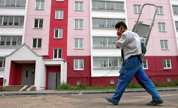 A man in casual attire carries a ladder over his shoulder as he walks by a brightly painted apartment complex, engaged in a phone conversation.
