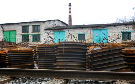 Rusting metal sheets are stacked near a railway track, with an old factory building and a tall smokestack visible in the background under a gray sky.