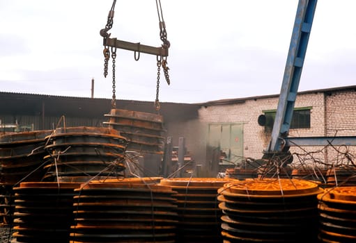 A crane hoists large, circular metal plates at an industrial site. Surrounding stacks of rusty plates indicate ongoing construction during overcast weather.