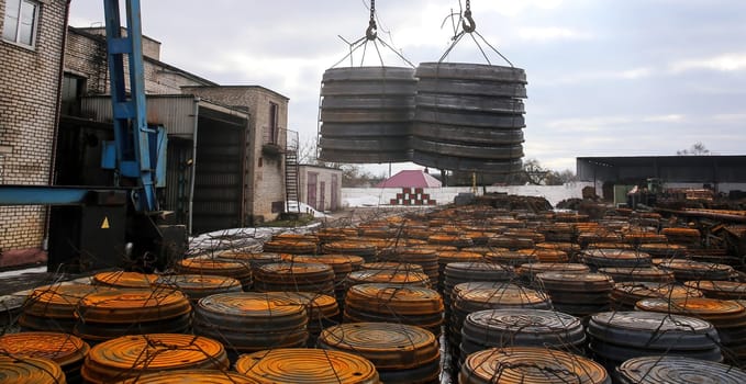A crane is actively lifting heavy metal coils at a storage facility, surrounded by stacks of similar coils on a chilly, overcast day. The environment highlights industrial operations.