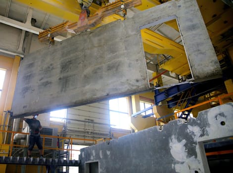 A construction worker is carefully positioning a large concrete wall panel using overhead cranes in a well-lit industrial area, showcasing teamwork in construction.