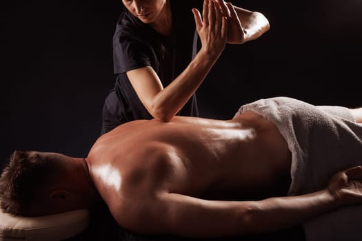 Girl gives back massage to muscular man on dark background close-up, beautiful relaxing male massage, recovery
