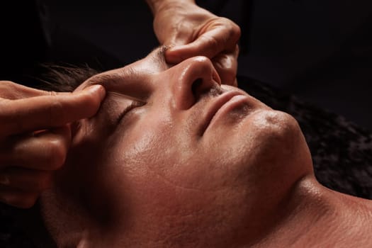 girl masseur doing facial massage to man on dark background close-up, facial skin rejuvenation, relaxation and meditation