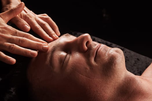 girl masseur doing facial massage to man on dark background close-up, facial skin rejuvenation, relaxation and meditation