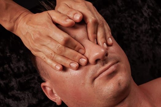 Close up of man receiving facial massage, masseur's hands massaging facial skin on dark background