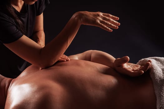 Girl gives back massage to muscular man on dark background close-up, beautiful relaxing male massage, recovery
