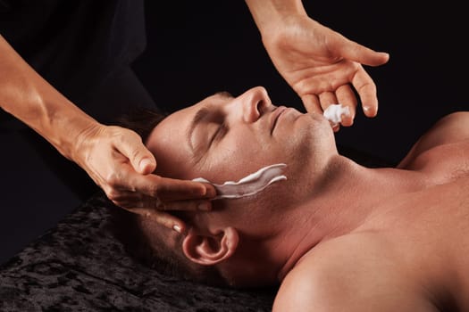 A man is getting a facial treatment at a spa. He is lying on a dark surface with his eyes closed, and the masseur is applying cream to his face with gentle hands. Facial massage concept