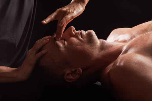 girl masseur doing facial massage to man on dark background close-up, facial skin rejuvenation, relaxation and meditation