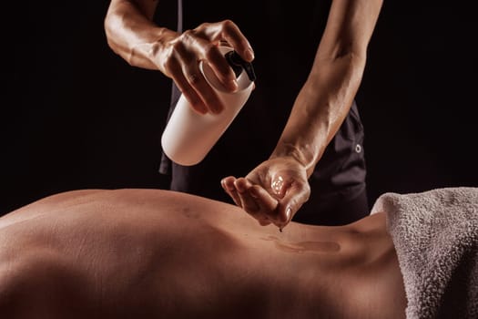 Girl giving back massage to muscular man on dark background close-up, pouring oil on body before massage