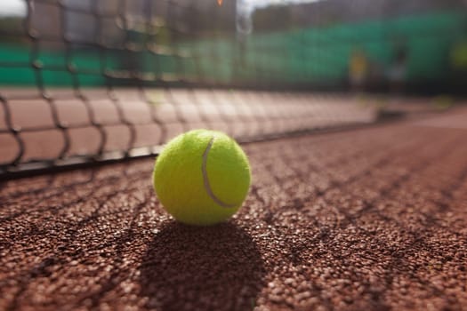 A close-up of a tennis ball resting on a clay court, with a net in the foreground and blurred players in the background