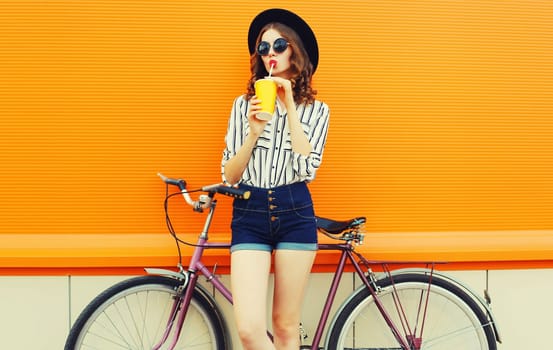 Summer colorful portrait of young woman drinking a juice with bicycle in city on orange background