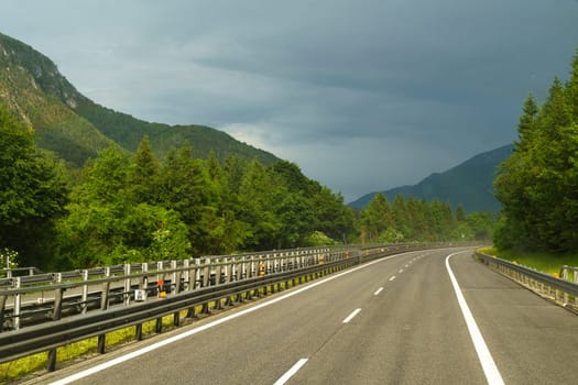A narrow highway meanders through vibrant green trees, flanked by majestic mountains, as dark clouds gather ominously overhead in the afternoon.