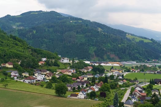 Nestled in a verdant valley, a quaint village features charming homes, winding roads, and lush greenery under a gentle blanket of clouds, creating a serene atmosphere.