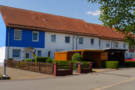 Waltershausen, Germany - June 11, 2023: A lovely residential structure in Waltershausen showcases a vibrant blue and white facade. The garden is neatly trimmed, with inviting hedges and shaded carports on a bright day.