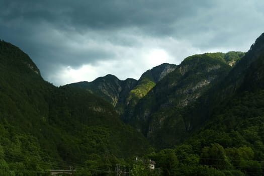 Dramatic cloud formations hover over towering mountains as the last light of day illuminates lush green valleys, creating a stunning contrast in the evening sky.