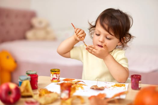 smiling little child girl painting her hands and autumn leaves. baby girl drawing hands in yellow paint