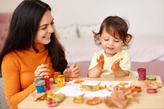 happy little child girl and mom spending good time painting hands and autumn yellow leaves together