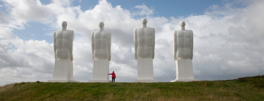 woman in re dcoat at the white Men statues, Esbjerg, Denmark. Four white men sitting looking at the sea near Esbjerg, Denmark. They are each 9 meters tall and made of white concrete.