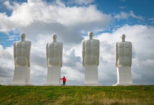 woman in re dcoat at the white Men statues, Esbjerg, Denmark. Four white men sitting looking at the sea near Esbjerg, Denmark. They are each 9 meters tall and made of white concrete.