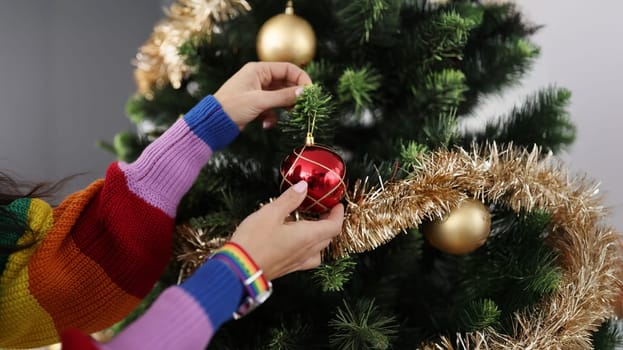 Close-up of woman in colourful sweater decorating christmas tree with red ball. Create cozy festive atmosphere indoors. Holiday, new year, decor concept