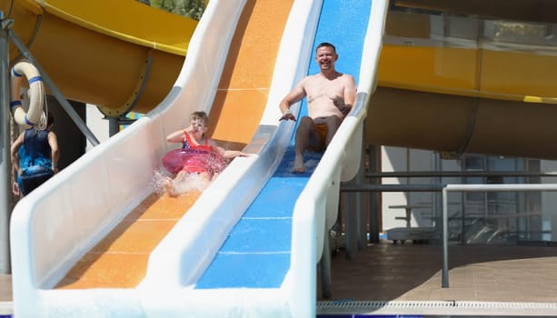 Portrait of father and daughter slide down hill on air mattress in water park. Smiling parent and child enjoy summer holiday. Summertime, leisure concept