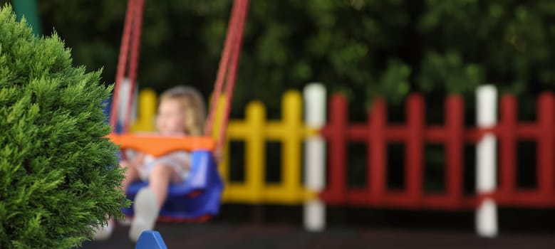 Close-up of green fluffy bush on playground child on swing in park alone. Little girl enjoy ride outdoors, place for children. Childhood, nature concept