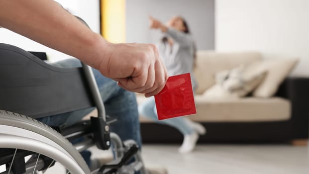 Close-up of man sit in wheelchair hold package of condom, woman laugh on couch pointing with finger. Disabled people, intimate life with disability concept