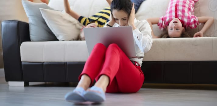 Low angle of mother try to concentrate and work while kids going crazy and jump. Female with pc close ears. Remote job, work from home, childhood concept