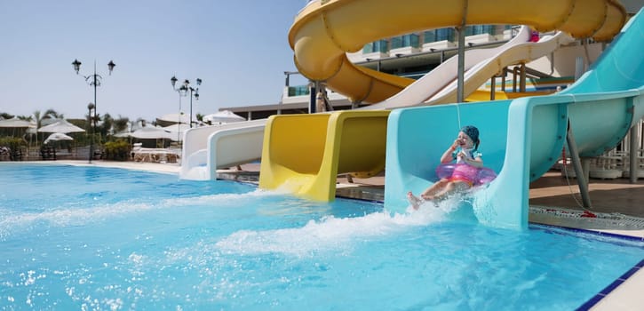 Portrait of happy little girl kid slide down hill on pink air mattress in water park. Smiling child enjoy summer vacation. Summertime, holiday, fun concept