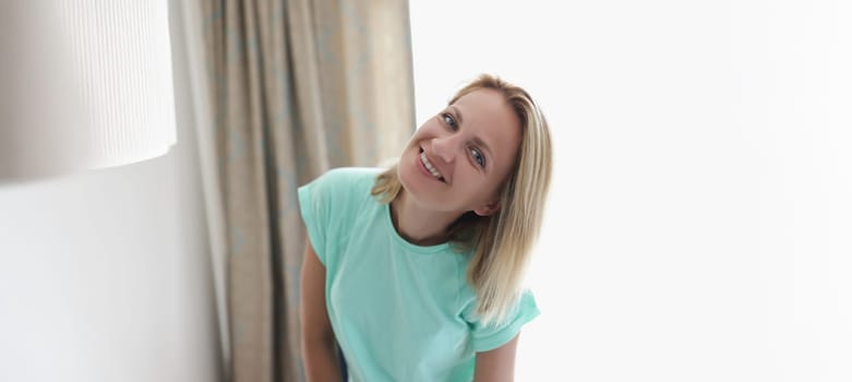 Portrait of happy smiling blonde young woman in apartment posing on curtain. Cheerful full of joy female at home. Modeling, relaxation, housewife concept