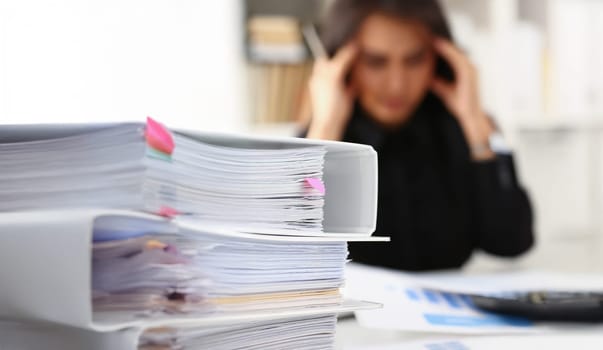 Close-up of tired and exhausted woman looks at documents touch head with hands. Huge pile of document folders on workplace. Headache, low energy concept