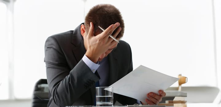 Close-up of adult man businessman hold paper sheet in hand and close face with palm. Wrong financial calculations led to failure. Business strategy concept