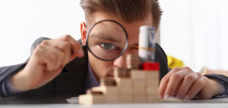 Close-up of businessman holds in hand black magnifying glass looks at tower of coins placed on wooden cubes. Financial development, invest, growth concept
