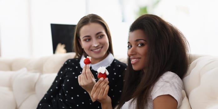 Portrait of smiling girls eating tasty fresh strawberry covered with whipped cream. Romantic date at home, best friends chilling. Lgbt, bff, lunch concept