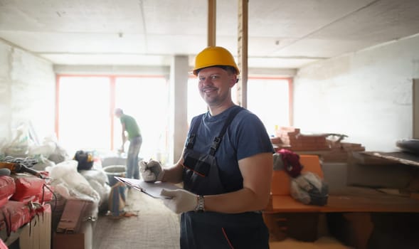 Portrait of smiling professional worker hold clipboard and list work materials. Joyful worker wear uniform and protective helmet. Construction site concept