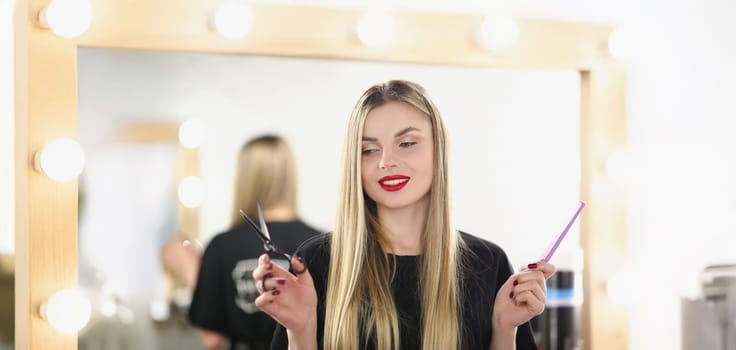 Portrait of young lovely professional master holding tools for work and posing. Qualified hairdresser hold scissors and comb. Beauty studio, barber concept