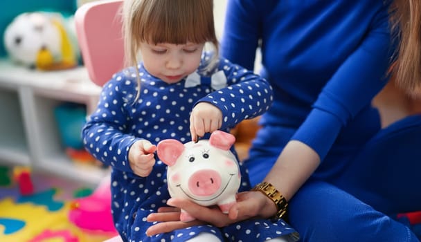 Portrait of cute little child putting coin cash into piggy bank container. Mother teach daughter to save up, save to buy toy. Childhood, investment concept
