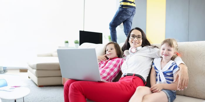 Portrait of happy woman posing with her kids on couch while working on laptop. Mother work from home, earn money for children. Parenthood, weekend concept
