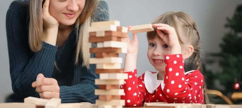 Portrait of pretty little kid pulling wooden block out of tower and smile. Mother join daughter in funny board game at home. Parenthood, childhood concept