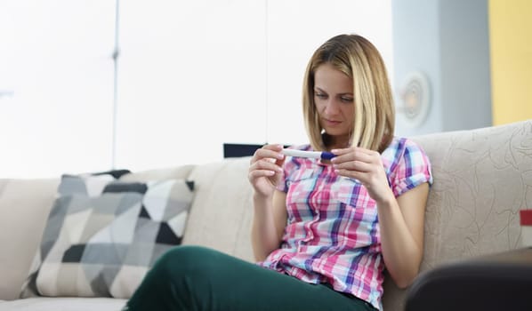 Portrait of young woman hold pregnancy test, happy female got big news. Blonde woman sit on sofa at home. Pregnancy, new period, life change, baby concept