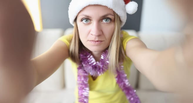 Portrait of beautiful serious blonde young woman posing on camera in santa claus hat. Celebrating new year alone at home. Holiday, christmas mood concept