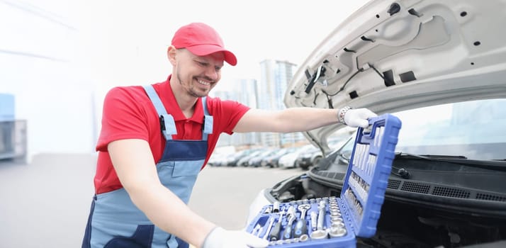 Portrait of automotive technician smiling and open kit full of equipments. Open car hood and need to fix problem of automobile. Car maintenance concept