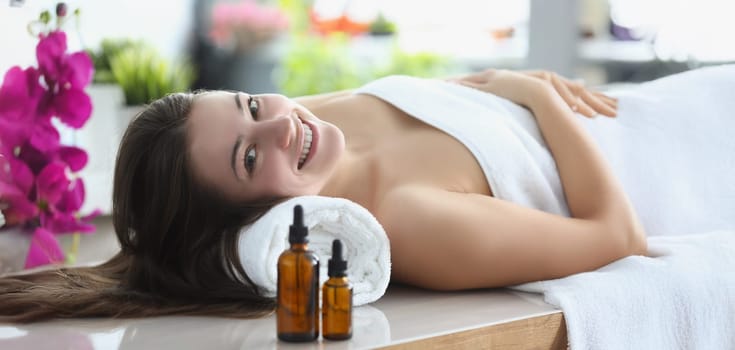 Portrait of smiling happy woman laying covered with towels in luxury spa centre. Rested female surrounded with beauty and smelly oils. Self care concept