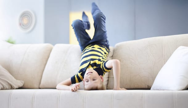 Portrait of young boy child standing on head on beige sofa going crazy at home on holiday. Alone at home, go nuts, lots of energy. Childhood, sport concept