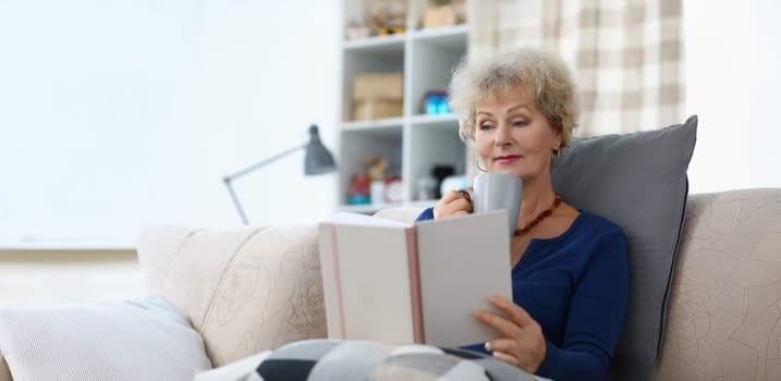 Portrait of old lady reading book and drinking cup of coffee being comfy on sofa at home. Attractive senior woman relaxing on weekend. Spare time concept