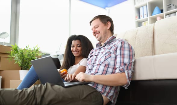 Portrait of friends sitting on carpet floor watching movie on laptop and laughing. Latino american woman eat apple. Spare time, friendship, leisure concept