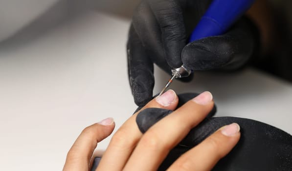 Close-up of nail master using tool to clean female clients nails in beauty studio. Worker wear protective black gloves for work. Care and beauty concept