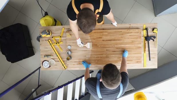 Top view of coworkers covering wooden plank with shiny polish creative process in workshop. Male carpenters finish masterpiece in studio. Design concept