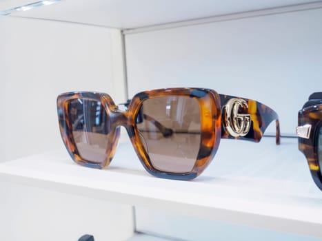 Brown tortoiseshell sunglasses with gold logo on the arm are displayed on a white shelf under bright lights