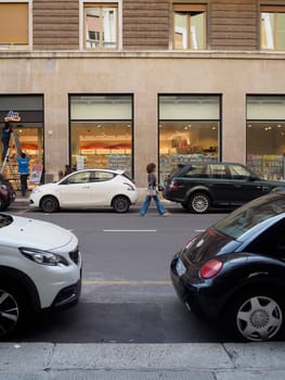Cremona, Italy August 31st 2024 A woman is walking in a european city street with parked cars on the side and workers setting up a shop